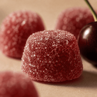 Macro close-up of sugar-coated cherry THC + CBD gumdrops showing sparkling sugar crystal texture and rich translucent cherry color on a warm neutral background.